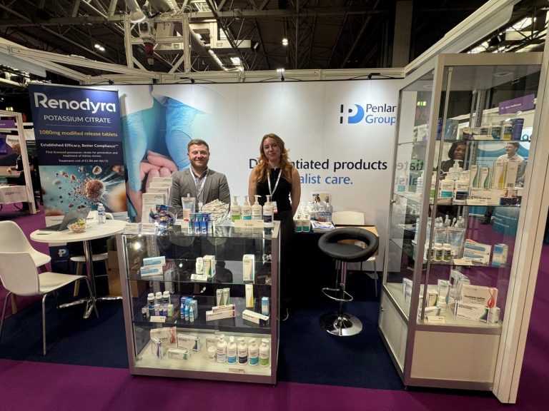 Two people standing behind a healthcare products exhibition stand at a trade show. The stand features Penlan Group branding, a large Renodyra Potassium Citrate banner, display cabinets filled with medical and skincare products, and promotional materials arranged on counters. The exhibition hall has purple carpeting and other healthcare exhibitors visible in the background.